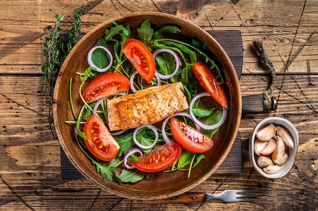 Salada de bife de salmão com folhas verdes de rúcula, abacate e tomate em uma placa de madeira. Fundo de madeira. Vista do topo.