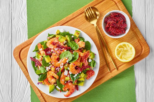 Salada de camarão com abacate e espinafre com tomate seco, queijo ralado e cebola roxa em um prato sobre uma placa de madeira