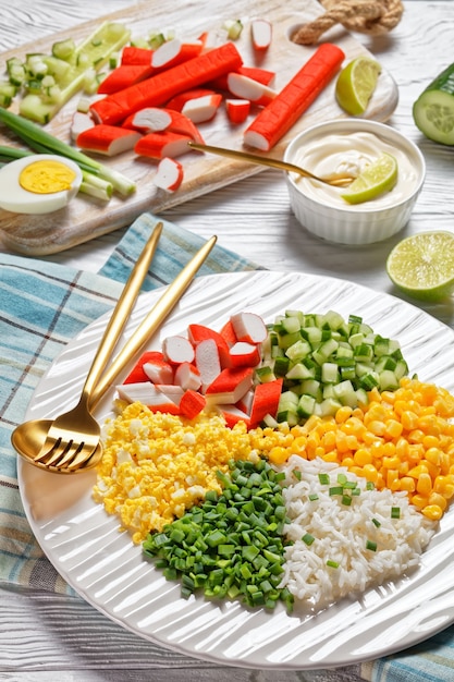 Salada de caranguejo de surimi com milho enlatado, pepino, ovo cozido de cebolinha, arroz de jasmim dividido em setores em um prato branco sobre fundo branco de madeira, vista de cima, close-up