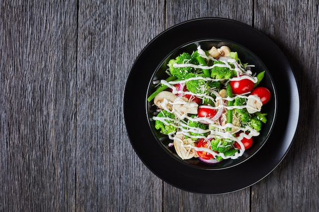 Salada de florzinhas de brócolis, feijão verde cozido no vapor, champignon, tomate coberto com queijo parmesão ralado e molho de iogurte em uma tigela preta, vista da paisagem de cima, planta plana, espaço livre