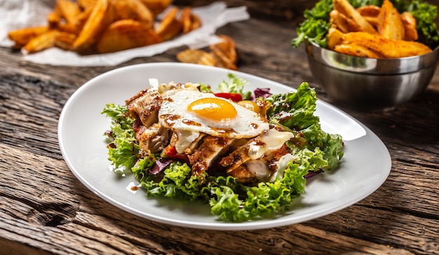 Salada de frango com ovo estrelado e molho em prato branco e madeira rústica, com rodelas de batata a acompanhar.