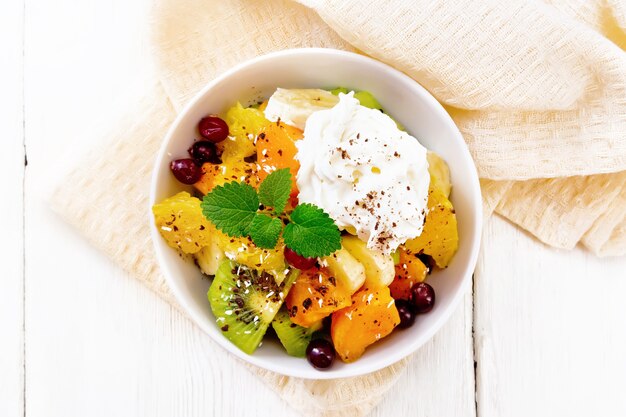 Salada de frutas com banana, laranja, kiwi, cranberries e abóbora assada, chantilly, polvilhada com chocolate e coco com hortelã em uma tigela em um guardanapo no fundo da placa de madeira de cima