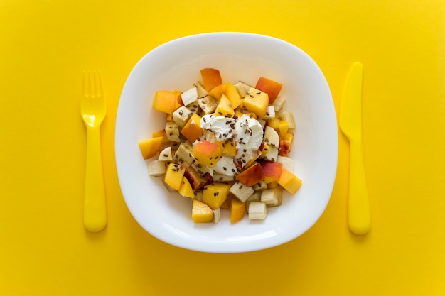 Salada de frutas de nutrição saudável com queijo macio e sementes de linho em chapa branca sobre fundo amarelo com garfo e faca amarela.