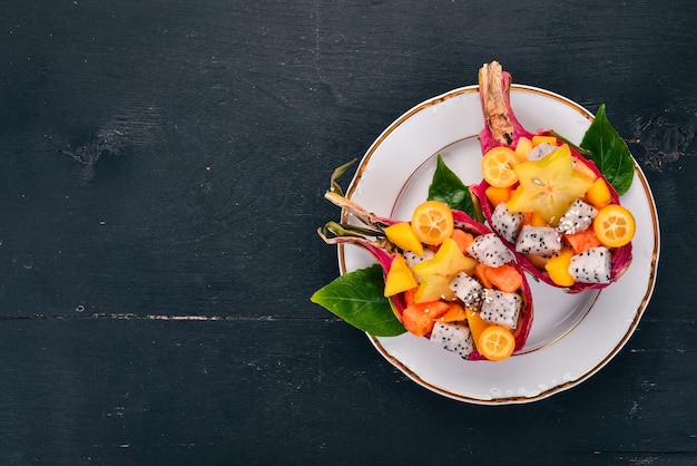 Salada de frutas tropicais servida em metade de uma fruta do dragão mamão rambutan tamarindo cacto manga de frutas em um fundo de madeira vista superior