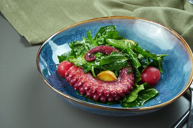 Salada de luxo com polvo grelhado, tomate e espinafre em um prato azul em uma mesa de madeira. Foco seletivo
