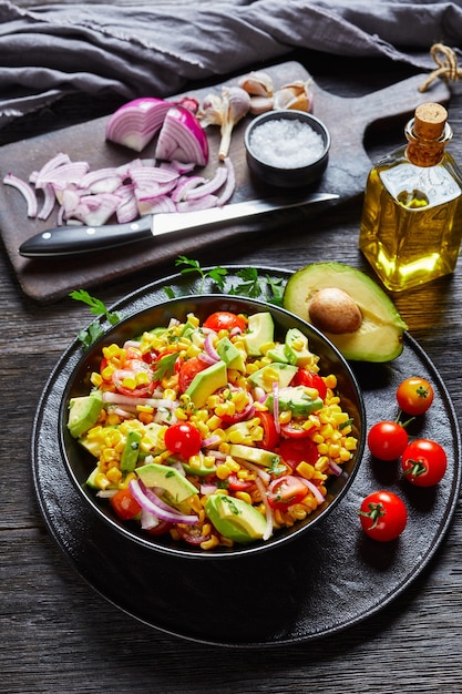 Salada de milho doce com abacate, fatias de cebola roxa e tomates em uma tigela preta sobre uma mesa de madeira escura com ingredientes na velha tábua escura, cozinha mexicana, visão vertical de cima