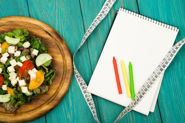 Salada de plano de dieta de fita métrica de legumes frescos e caderno em branco em uma mesa de madeira branca