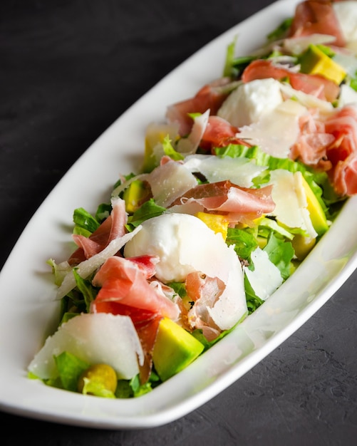 Salada de queijo de búfala e presunto serrano, acompanhada de abacate. Fundo preto. Prato branco.