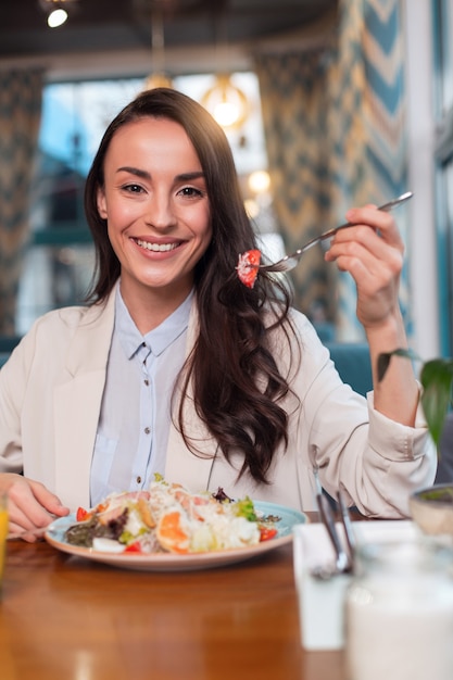 Salada deliciosa. Mulher alegre irresistível e feliz levantando o garfo enquanto olha para a câmera e se senta à mesa