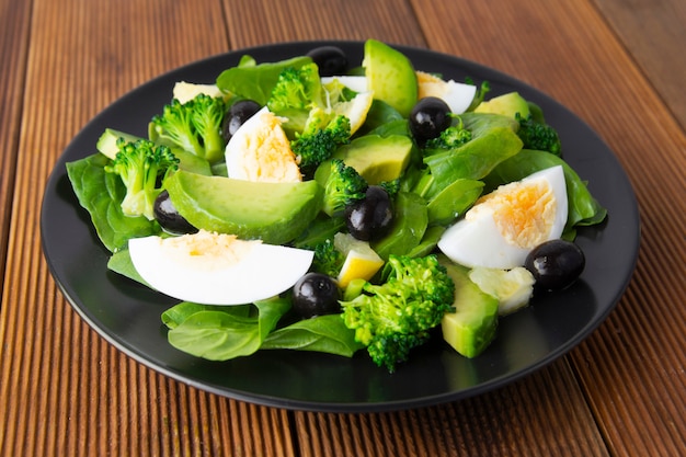 Salada do abacate com brócolis, espinafres, azeitonas e ovos cozidos na placa preta, tabela de madeira.