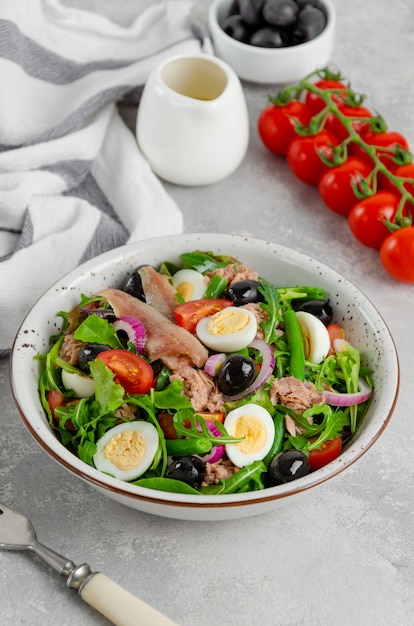 Salada francesa Nicoise com atum, ovo, feijão verde, tomate, azeitonas, alface, cebola e anchovas em um fundo cinza de concreto.