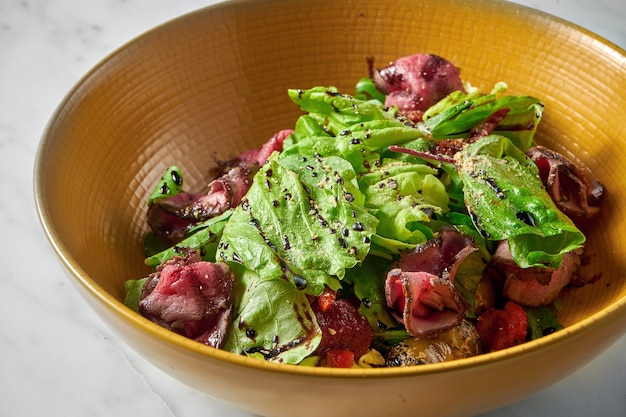 Salada fresca com fatias de rosbife mal passado, com tomate cereja em uma tigela amarela na mesa de mármore branco