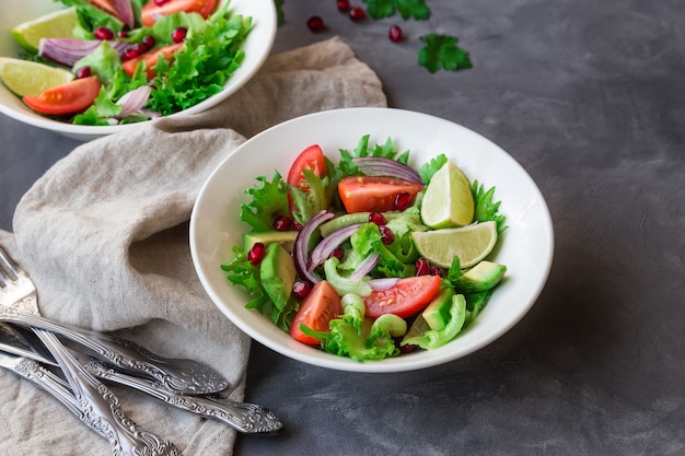 Salada fresca e saudável com tomate, abacate e romã em tigelas em concreto cinza.