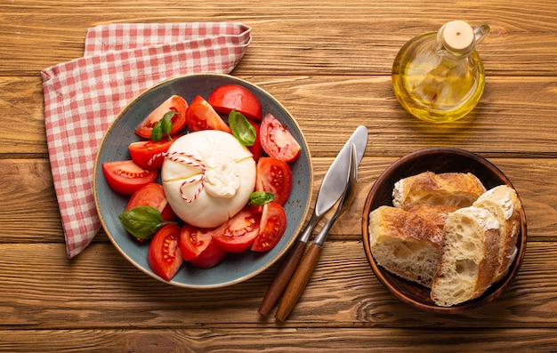 Salada italiana com tomate fresco, queijo burrata, manjericão servido em prato de cerâmica com pão de ciabatta e azeite em garrafa de vidro em fundo de madeira rústico de cima, lanche saudável ou almoço