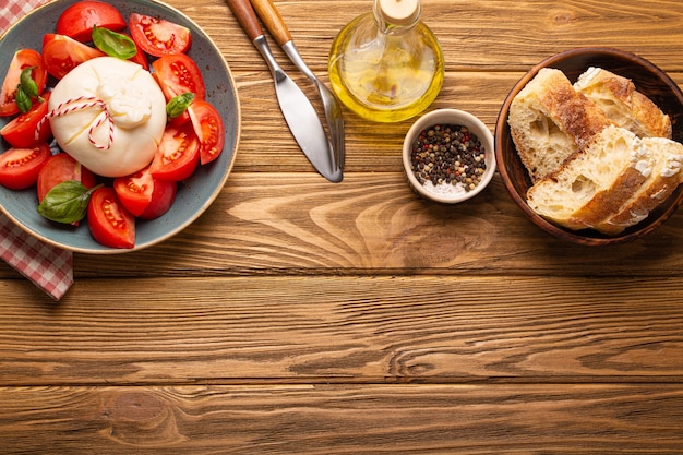 Salada italiana com tomates frescos, queijo burrata, manjericão servido em prato de cerâmica com pão ciabatta e azeite em garrafa de vidro em fundo de madeira rústico de cima com espaço para texto