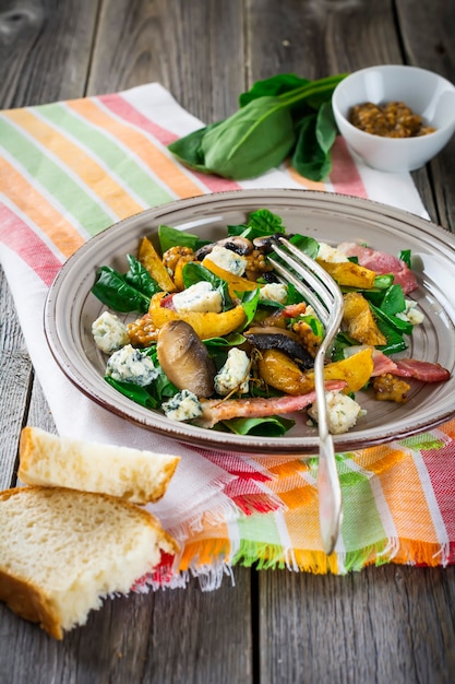 Salada quente de outono com batatas, cogumelos, bacon, espinafre, molho de mostarda e queijo em um guardanapo de laranja. Superfície de madeira velha. Foco seletivo.