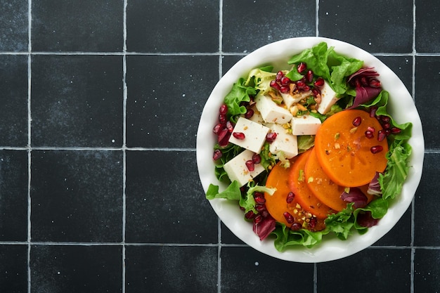 Salada Salada verde fresca com queijo feta, caqui, romã e pistache, em prato branco, em velhos azulejos pretos Idéia para um inverno saudável e delicioso Salada de Natal Alimentação saudável e equilibrada Vista superior