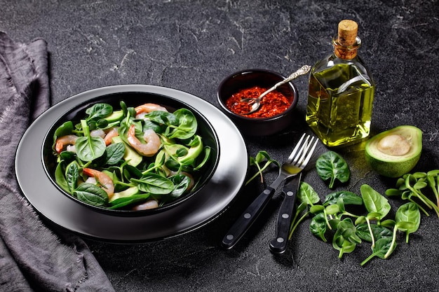 Salada saudável de camarão abacate com folhas de espinafre, brotos de girassol frescos, molho de azeite e flocos de pimenta, servidos em um prato em um fundo escuro com talheres, vista de cima, close-up