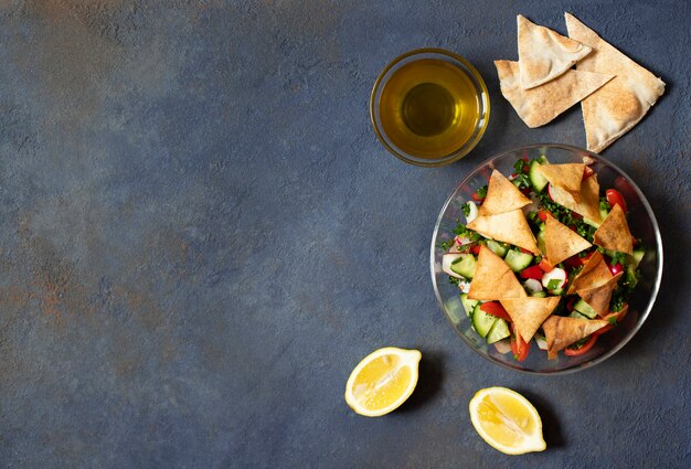 Salada tradicional de Fattoush com legumes e pão pita. Levantine, árabe, culinária do Oriente Médio. Servido em um prato de vidro com limão, pão árabe e azeite. Fundo escuro. Vista do topo. Espaço para texto