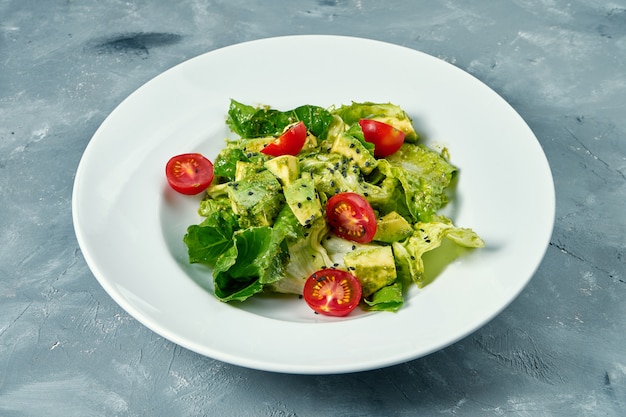 Salada vegetariana com abacate, alface e tomate cereja em um prato branco sobre uma superfície de concreto