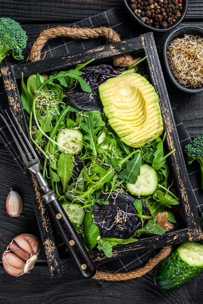 Salada vegetariana verde com mistura de folhas de salada, abacate e vegetais. Fundo de madeira preto. Vista do topo.