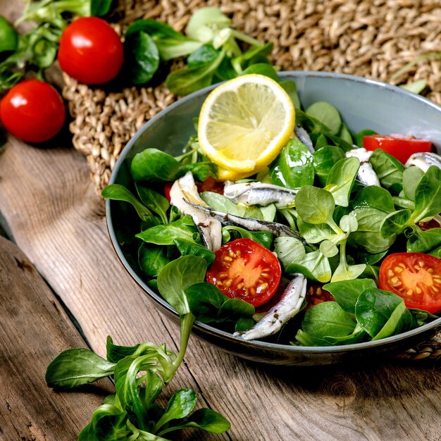 Salada verde com anchovas em conserva ou filé de sardinha e tomate cereja, servido em uma tigela azul com limão e azeite no guardanapo de palha sobre a velha mesa de madeira. Imagem quadrada