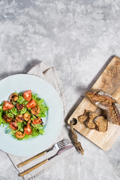 Salada verde com camarão grelhado e placa de corte de madeira com pão.