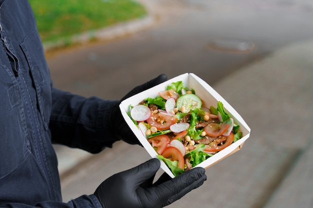 Salada verde com vitela e vagantes. entrega segura com máscara e luvas médicas pretas em quarentena. salada em caixa térmica ecológica.