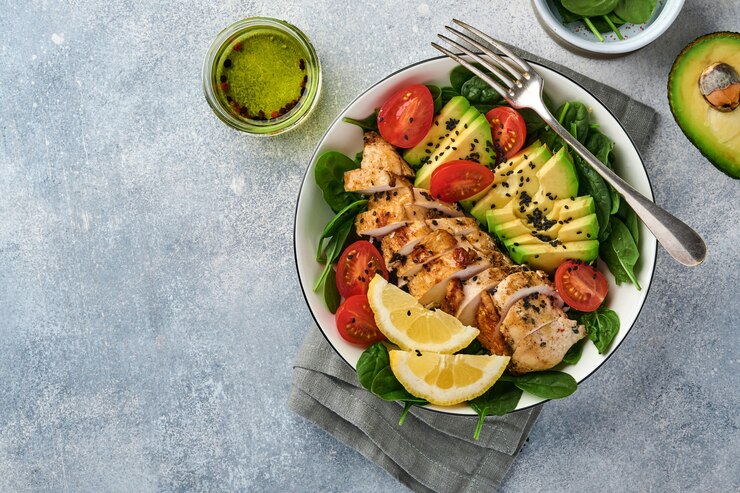 Salada verde fresca com filé de frango grelhado, espinafre, tomate, abacate, limão e sementes de gergelim preto com azeite em uma tigela branca sobre fundo claro de ardósia. Conceito de dieta de nutrição. Vista do topo