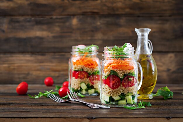 Saladas com quinoa, rúcula, rabanete, tomate e pepino em potes de vidro na mesa de madeira. Comida saudável, dieta, desintoxicação e conceito vegetariano