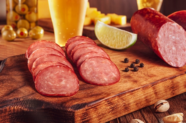 Salami ahumado en rodajas sobre tabla de cortar con vaso de cerveza, cubos de queso, aceitunas y pistachos