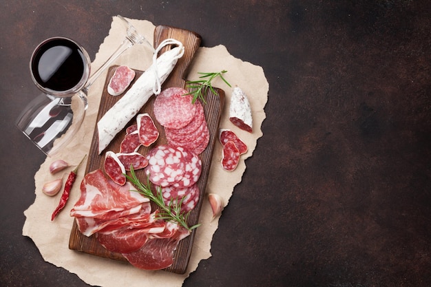 Salami, jamón rebanado, salchicha, prosciutto, tocino y copa de vino tinto. Plato de antipasto de carne en una mesa de piedra. Vista superior con espacio de copia