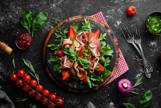 Salat aus Arugula-Prosciutto-Erdbeeren und Kapern auf einer schwarzen Steinplatte Italienische Küche
