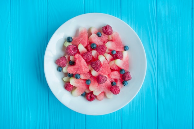 Salat aus Früchten und Beeren. Leichter herzförmiger Wassermelonensalat, Himbeeren, Blaubeeren in einer weißen Platte auf einem blauen hölzernen Hintergrund. Vegetarisches Gericht. Sommergericht. Gesunde Lebensmittelbilanz.