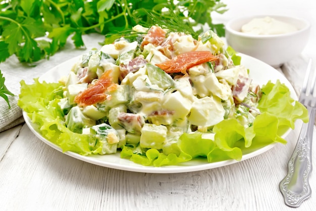 Salat aus Lachs, Gurke, Eiern und Avocado, angerichtet mit Mayonnaise auf Salatblättern in einem Teller, Handtuch, Dill, Petersilie und Gabel auf hellem Holzbretthintergrund