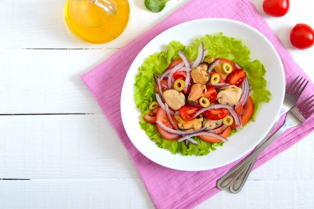 Salat aus marinierten Muscheln, frischen Tomaten, roten Zwiebeln, Oliven in einer Schüssel auf einem weißen Holztisch. Draufsicht.