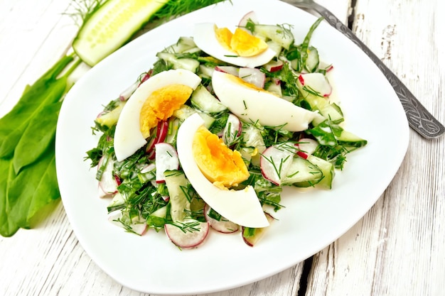 Salat aus Radieschen, Gurke, Sauerampfergrün und Eiern, gekleidet mit Mayonnaise und Sauerrahm in einem weißen Teller auf dem Hintergrund eines Holzbretts