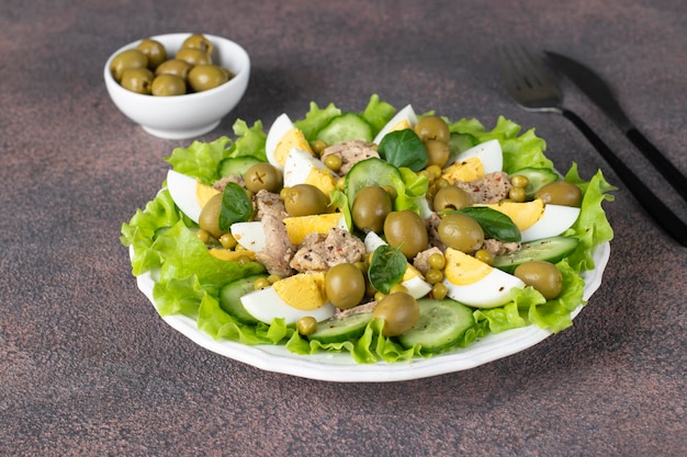 Salat mit Dorschleber, Ei, Gurke, Oliven und Erbsen, serviert auf grünen Salatblättern auf weißem Teller auf braunem Tisch