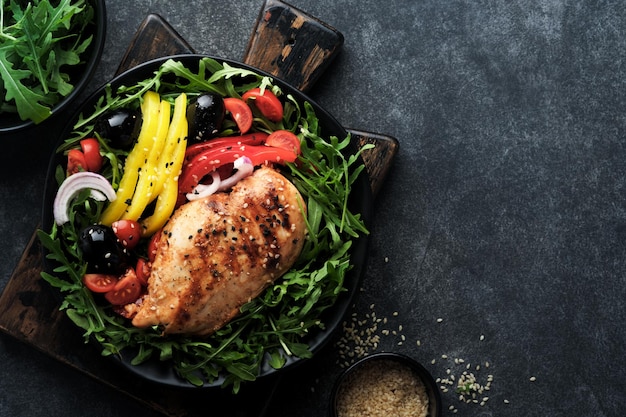 Salat mit gegrilltem Hähnchenbrustfilet mit frischen Rucola-Tomaten und Oliven auf schwarzem Teller auf altem dunklen Hintergrund Hühnerfleisch mit Salat ketogenes Mittagessen gesunde Ernährung Draufsicht