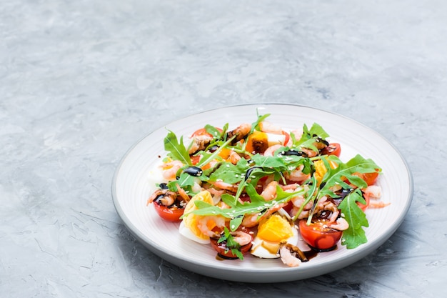 Salat mit Kirschtomaten, Eiern, gekochten Garnelen, Rucola, Sesamsamen und Balsamico-Sauce in einer Platte auf dem Tisch