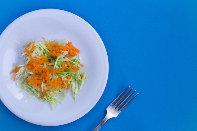 Salat mit Kohl und Karotten in der weißen Platte auf blauem Hintergrund. Gesunde Lebensmittelzutaten. Ansicht von oben. Platz kopieren.