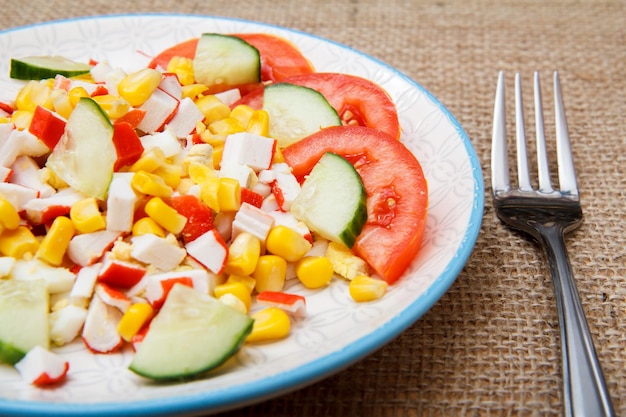 Salat mit Krabbenstäbchen, gekochtem Mais und frisch geschnittenen Tomaten und Gurken auf Teller mit Gabel auf Sackleinen