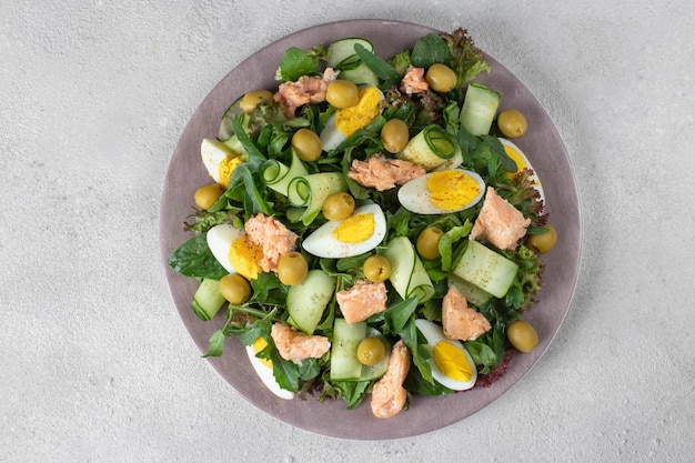 Salat mit Lachs-Ei-Gurken-Oliven und Rucola auf runder Platte auf hellgrauem Hintergrund