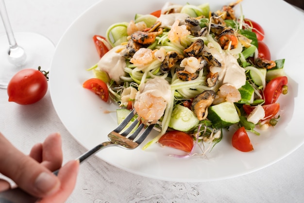 Salat mit Muscheln, Garnelen, Tomaten, Kirschen, Gurken, Zucchini-Nudeln, Mikrogrün, Joghurtsauce auf weißem Teller auf Leuchttisch.