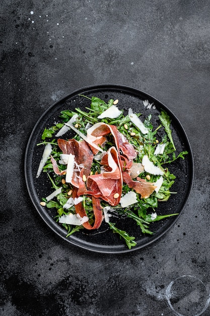 Salat mit Parma, Schinken, Rucola und Parmesan. Schwarzer Hintergrund, Draufsicht, Platz für Text
