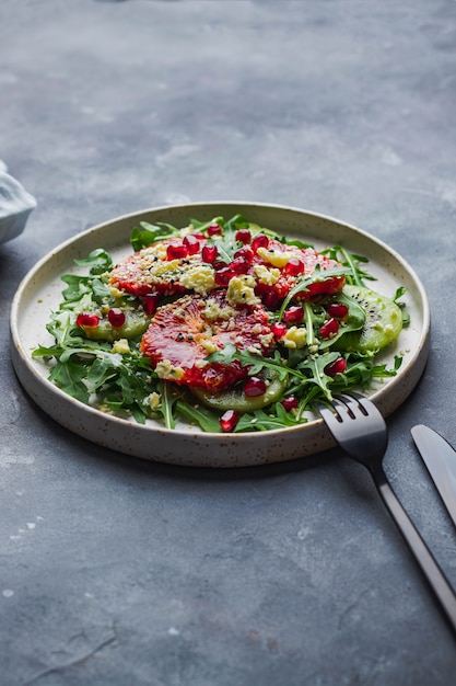 Salat mit Rucola, Kiwi, Blutorange, Meersame, Kümmel, Granat, dor blau auf blauer Steinmauer mit schwarzer Gabel und Messer angebraten. Konzept für gesunde Ernährung. Draufsicht mit Copyspace