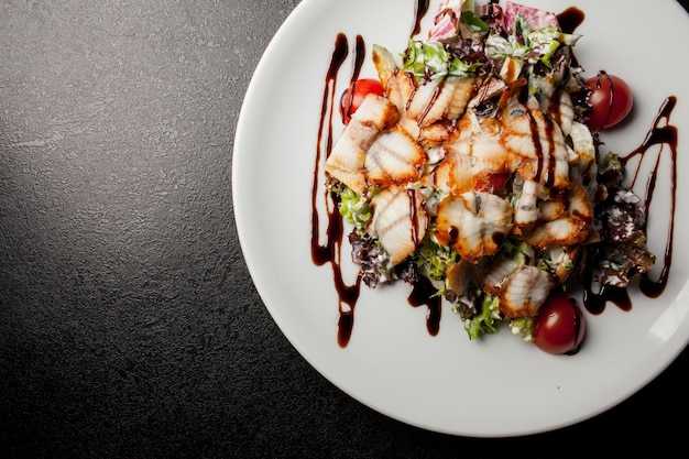 Salat mit Tintenfisch, Salat, Kohl, Kirschtomaten und Sauce. Salat mit Tintenfischscheiben auf einem weißen Teller auf schwarzem Hintergrund. Foto Nahaufnahme, Weichzeichner. Foto für Rezeptbuch, Restaurantkarte