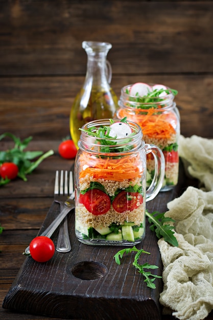 Salate mit Quinoa, Rucola, Radieschen, Tomaten und Gurken in Gläsern auf Holztisch. Gesundes Essen, Ernährung, Entgiftung und vegetarisches Konzept