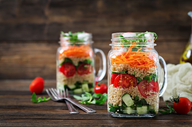 Salate mit Quinoa, Rucola, Radieschen, Tomaten und Gurken in Gläsern auf Holztisch. Gesundes Essen, Ernährung, Entgiftung und vegetarisches Konzept