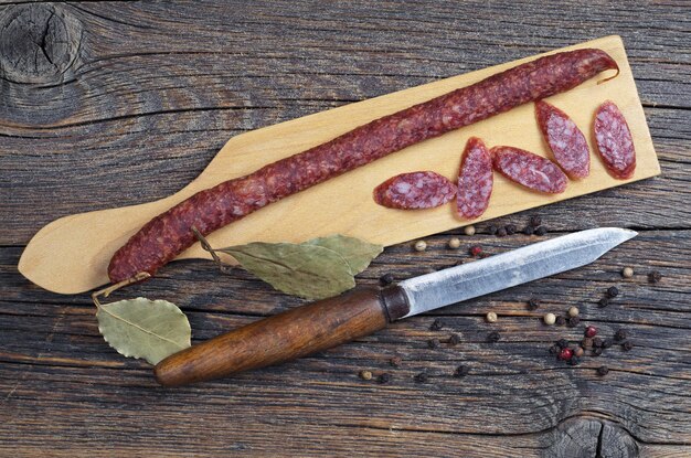 Salchicha ahumada delgada, cuchillo viejo, pimienta y hoja de laurel en una tabla de corte ubicada sobre un fondo de madera, vista superior